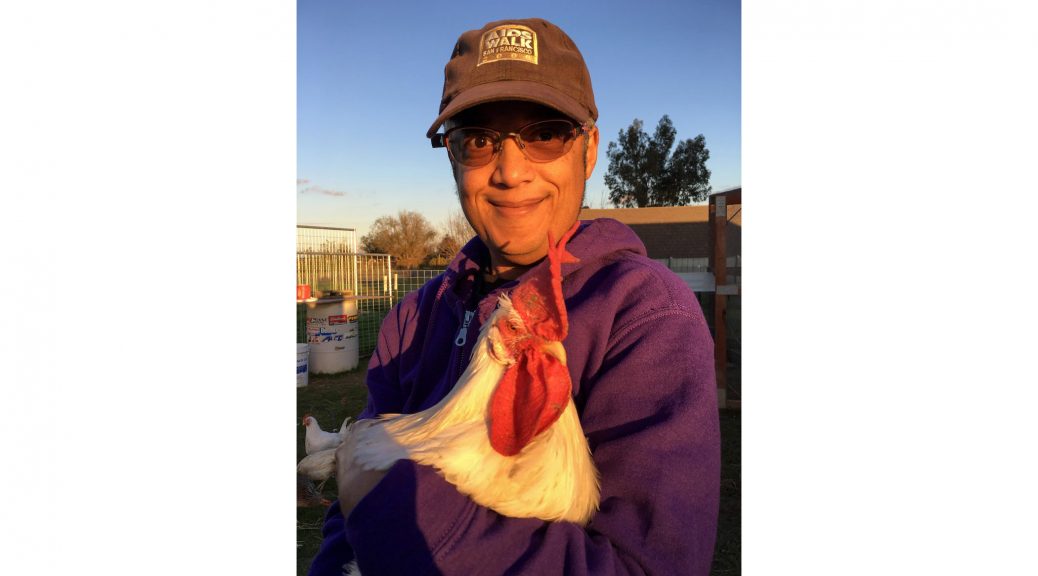 The author poses holding Ricky, a rooster, at PreetiRang Sanctuary. Pax is wearing a purple hoodie, tinted eyeglasses, and a faded black AIDS Walk San Francisco 2000 baseball cap. Photo by Ziggy, November 2016.