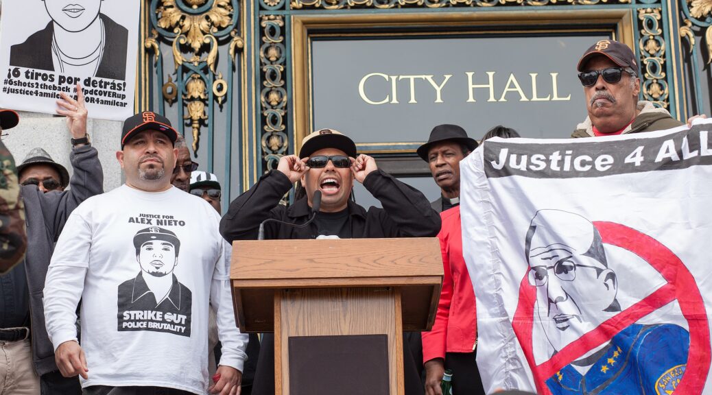Activist Benjamin Bac Sierra speaks at a podium on the steps of San Francisco City Hall. Next to him, another activist wears a shirt reading “Justice for Alex Nieto — Strike Out Police Brutality”. Another holds a banner partially reading “Justice 4 All”, with an image of the police chief crossed out in red. Another holds a sign reading in part, “¡6 tiros por detrás! #6shots2theback #sfpdCOVERup #justice4amilcar”.