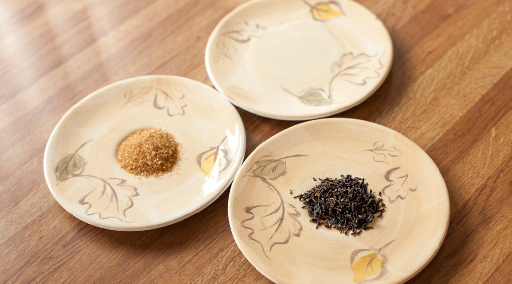 Three saucers on a wooden table containing small amounts of black tea leaves, turbinado sugar, and soy milk, respectively.