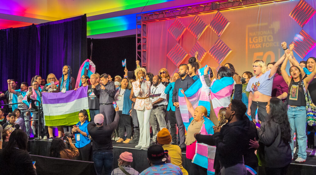 Angelica Ross speaks on an indoor stage, surrounded by people holding trans and non-binary pride flags and cheering.