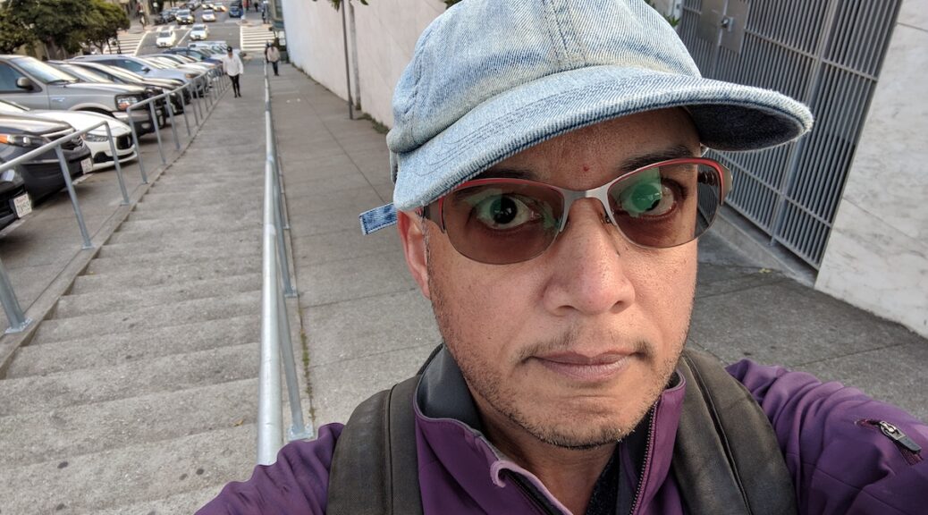 Selfie of Pax at the top of a steep outdoor staircase. They are wearing a blue denim cap, red-rimmed tinted glasses, and a purple jacket.