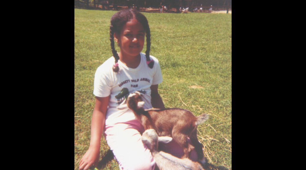 Pax at age 10 (summer of 1980), with their hair in two braids and wearing a Bassett Wild Animal Farm T-shirt, sitting in the grass and being happily nibbled on by baby goats.