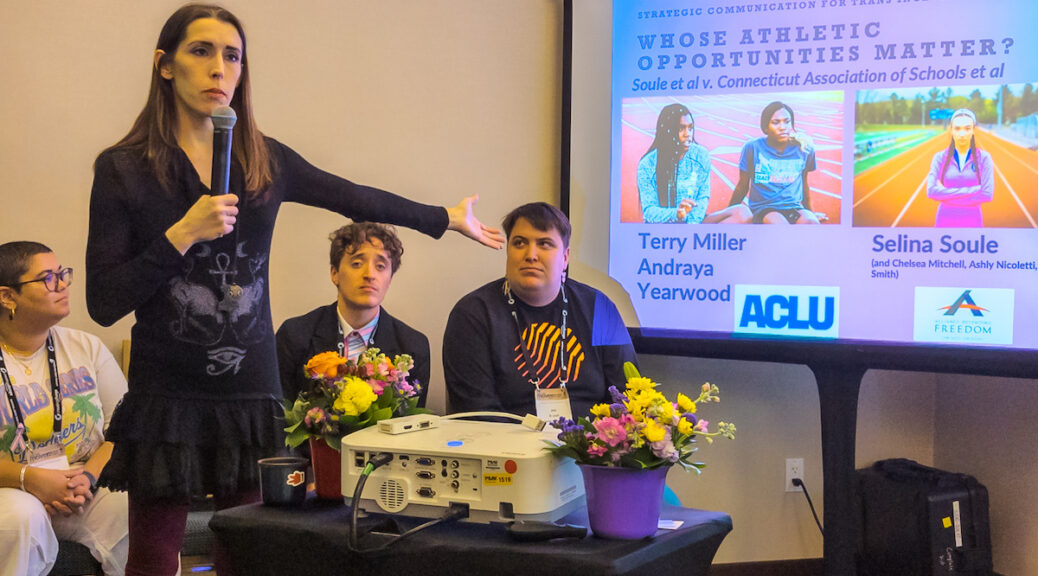 Trans advocate Kayley Whalen stands and speaks into a microphone while giving a presentation on trans athletes, while three seated panelists look on. The slide on the screen has text reading “Strategic Communication for Trans Inclusion in Sports”, “Whose Athletic Opportunities Matter?” “Soule et al. v. Connecticut Association of Schools et al”, a photo of athletes Terry Miller and Andraya Yearwood, a photo of athlete Selina Soule, and the logos of the ACLU and Alliance Defending Freedom.