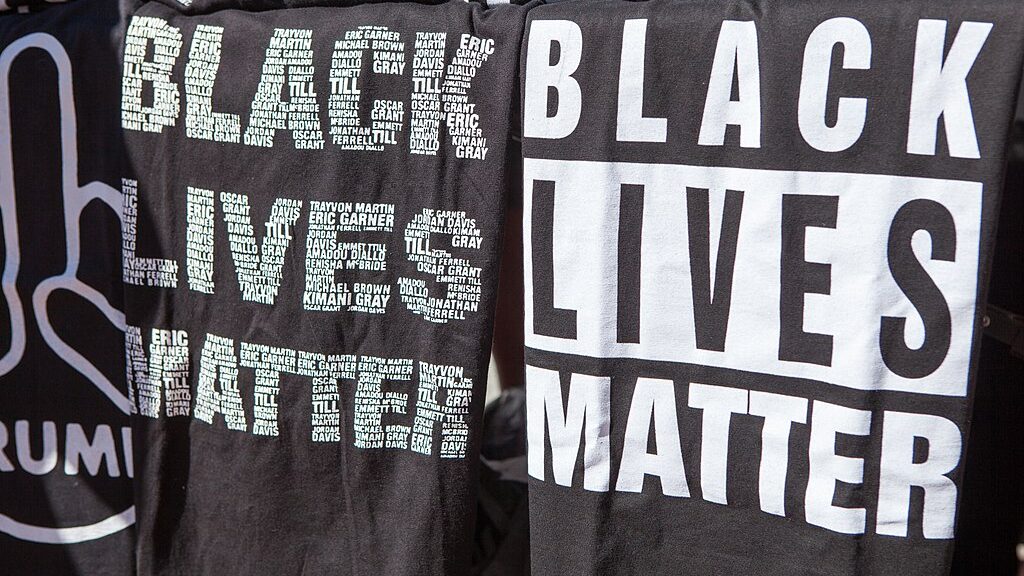 Two black and white T-shirts reading “Black Lives Matter”. On one of the shirts, the letters in the slogan are made out of the names of Black victims.