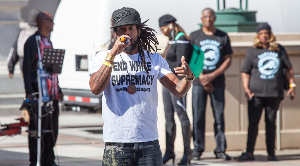 A Black man with long locs raps into a microphone outdoors, while wearing a black ball cap reading “Louder” and a white T-shirt reading “END WHITE SUPREMACY”.
