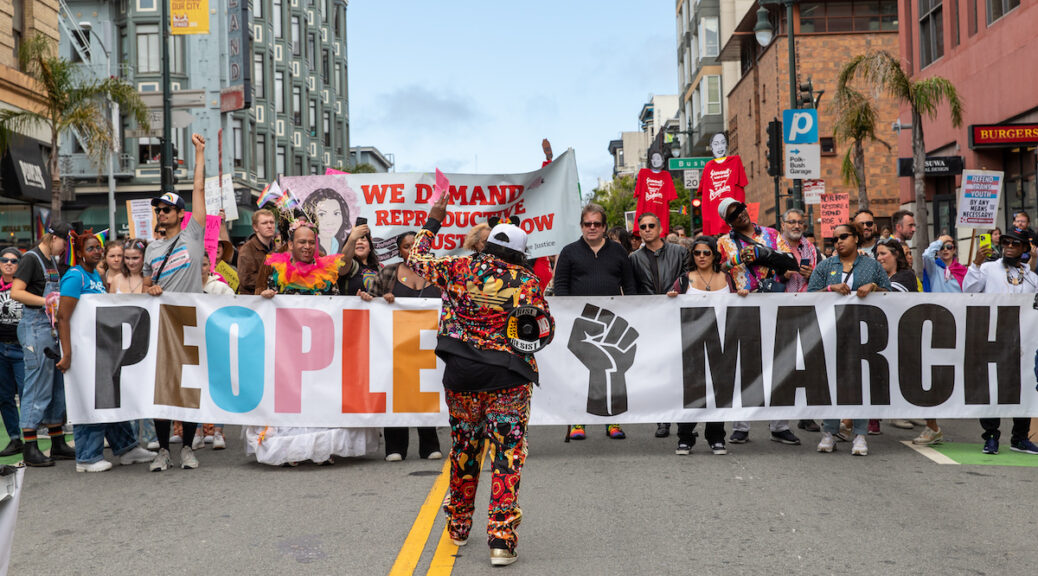 A crowd of people stands on Polk Street, San Francisco, behind a banner reading “People’s March”. Event co-organizer Alex U. Inn stands in front with a megaphone.
