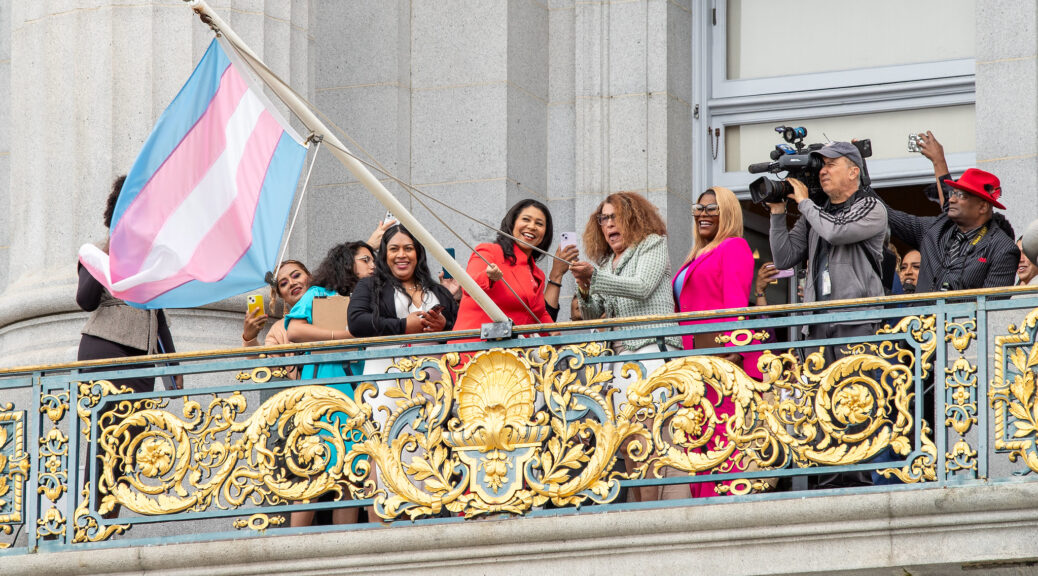 San Francisco Mayor London Breed and trans activist Donna Personna raise the trans pride flag outside City Hall, surrounded by guests and photographers on the mayor’s balcony.