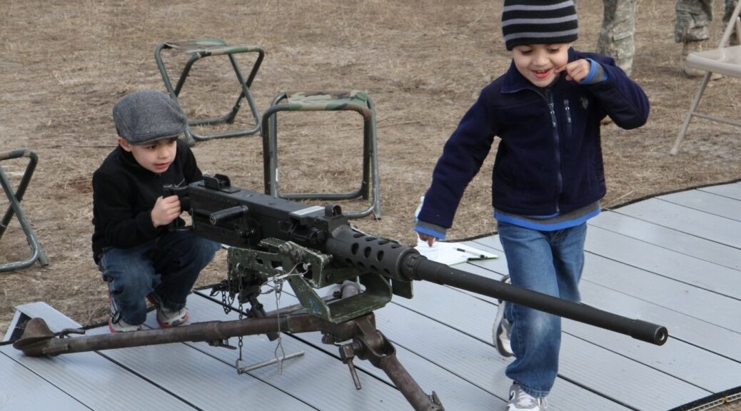 A young child interacts with an M2 machine gun on display outdoors while another watches.