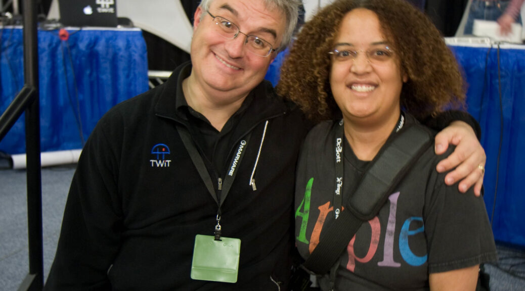 Image: Leo Laporte and Pax sit next to each other and smile for the camera. Both are wearing lanyards with badges. Leo has his arm around Pax, who is wearing an Apple T-shirt.