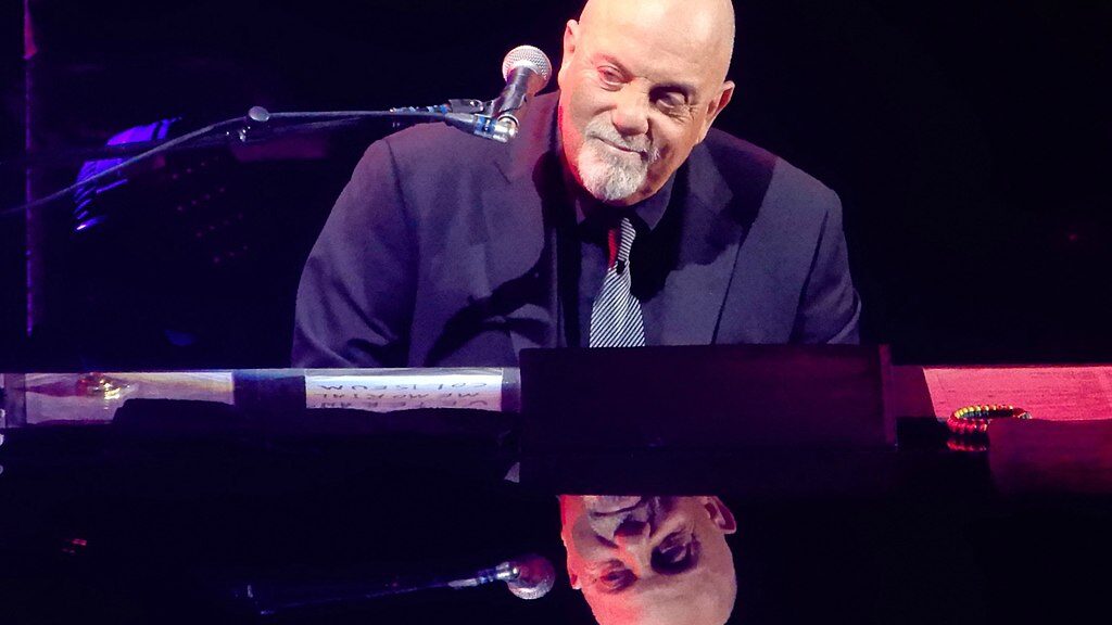 Billy Joel sitting a piano, smiling slightly and looking to the side. His face is reflected in the piano’s surface.