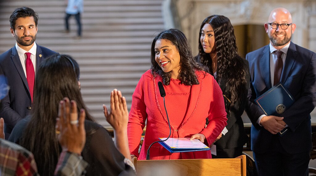 London Breed stands at a podium in San Francisco City Hall, surrounded by City officials.