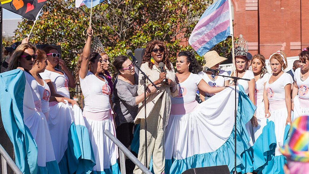 On an outdoor stage on a sunny day, Janetta Johnson speaks into a mic, surrounded by members of El/La Para TransLatinas, some holding Pride flags