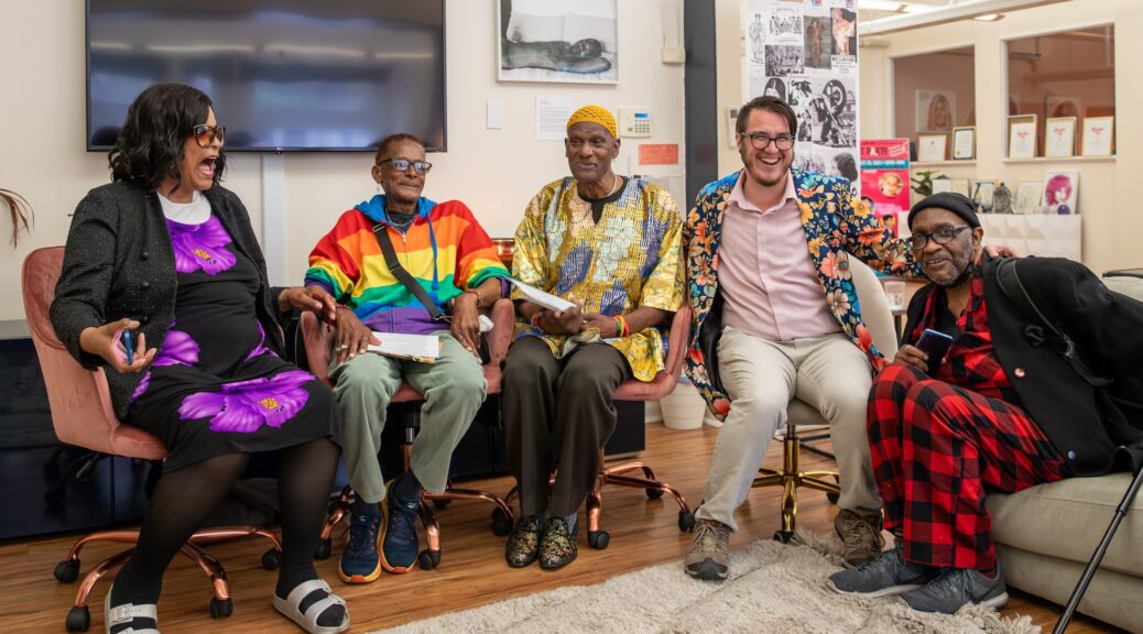 Five people - four Black, one white - wearing colorful clothing sit in chairs and on a sofa, laughing and smiling at the viewer.